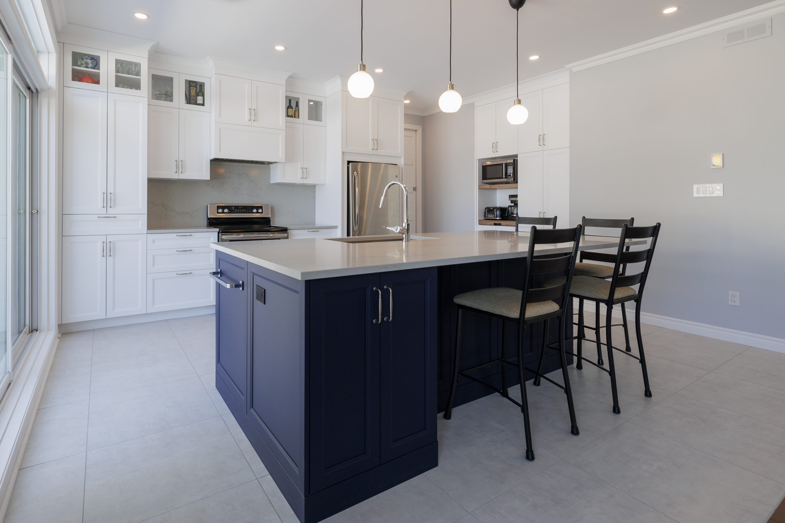 Modern kitchen with navy blue island featuring stools, round pendant lighting and clean white cabinets.